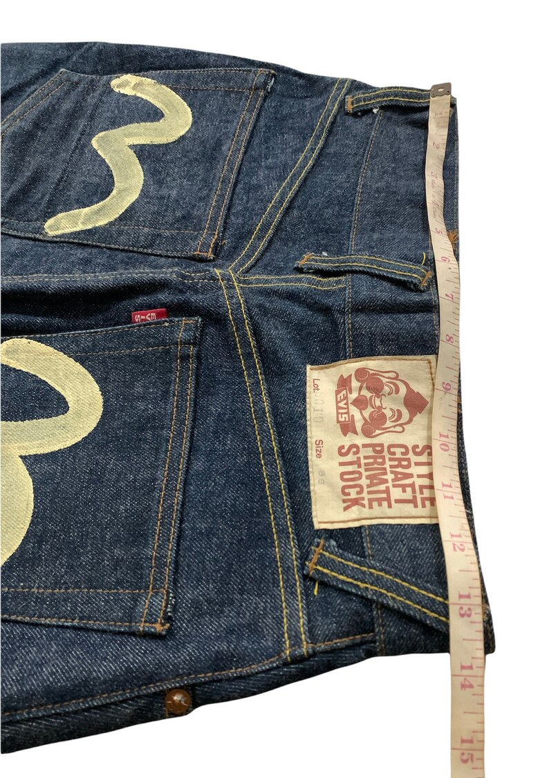 Pu&ograve; includere: Jeans in denim blu con un designo ricamato bianco sulle tasche posteriori. I jeans hanno una toppa in pelle marrone con il testo "Style Craft Private Stock EVIS" e "Lot. 010 Taglia 36".