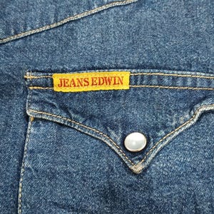 Pode incluir: Close-up de um bolso de camisa de ganga com uma etiqueta amarela que diz "JEANS EDWIN". O bolso tem uma aba decorativa com um &uacute;nico bot&atilde;o perolado. A camisa &eacute; de cor azul m&eacute;dia com costuras vis&iacute;veis.