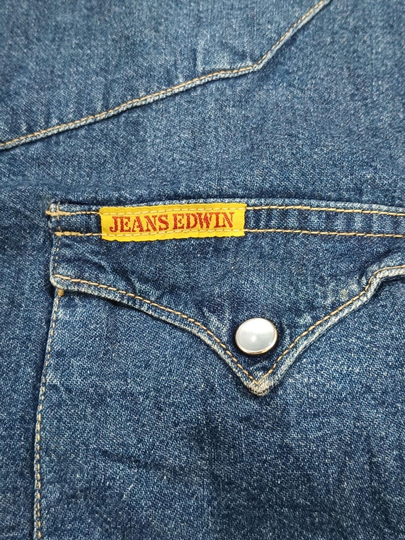 Pode incluir: Close-up de um bolso de camisa de ganga com uma etiqueta amarela que diz "JEANS EDWIN". O bolso tem uma aba decorativa com um &uacute;nico bot&atilde;o perolado. A camisa &eacute; de cor azul m&eacute;dia com costuras vis&iacute;veis.