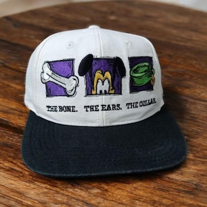 Puede incluir: Gorra de béisbol blanca con visera negra. La gorra presenta imágenes bordadas de un hueso, orejas y un collar, cada uno dentro de un cuadrado morado. Debajo de las imágenes está el texto "THE BONE. THE EARS. THE COLLAR."
