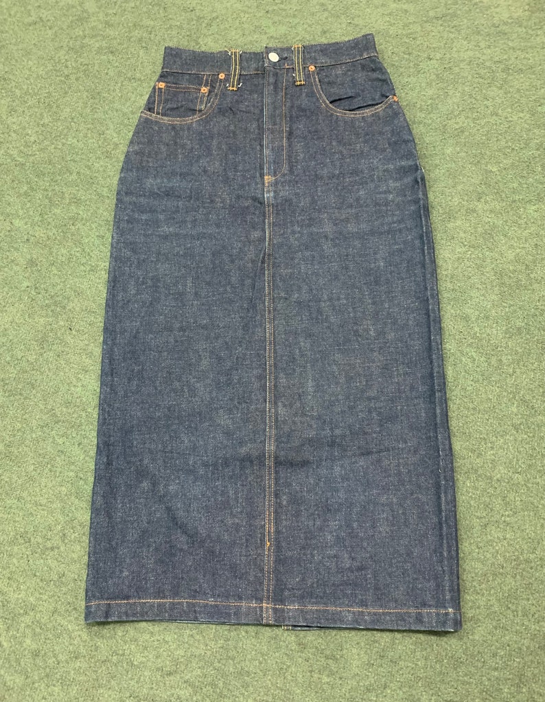 Pu&ograve; includere: Una gonna in denim blu scuro con vita alta e silhouette dritta e lunga. La gonna ha una chiusura con bottone e zip sul davanti e cinque tasche.