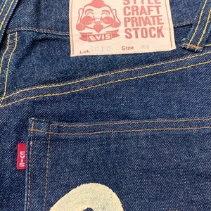Pu&ograve; includere: Un paio di jeans in denim blu scuro con una toppa rossa EVISU sulla tasca posteriore. La toppa presenta un personaggio dei cartoni animati sorridente e il testo "STYLE CRAFT PRIVATE STOCK". I jeans hanno anche un logo EVISU bianco sulla tasca posteriore. L'etichetta sul retro della cintura dice "Lot. 2010 Taglia 38".