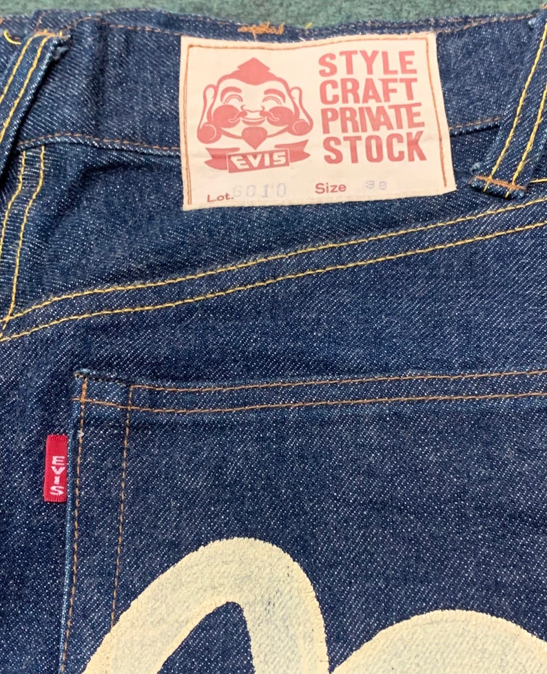 Pu&ograve; includere: Un paio di jeans in denim blu scuro con una toppa rossa EVISU sulla tasca posteriore. La toppa presenta un personaggio dei cartoni animati sorridente e il testo "STYLE CRAFT PRIVATE STOCK". I jeans hanno anche un logo EVISU bianco sulla tasca posteriore. L'etichetta sul retro della cintura dice "Lot. 2010 Taglia 38".