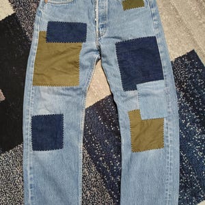 Può includere: Jeans in denim azzurro con chiusura a bottoni e diverse toppe rettangolari in verde oliva e blu navy scuro. Le toppe sono fissate con impunture bianche a vista che creano un contrasto sul tessuto in denim azzurro sbiadito.