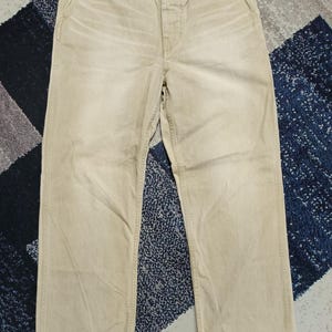 May include: Beige cotton trousers featuring a button fly closure, a waistband with belt loops, two side pockets, and a small coin pocket. The fabric has a slightly faded, worn-in appearance with visible stitching details along the seams and pockets.