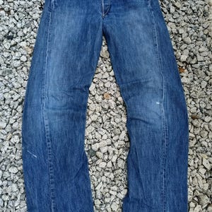 Puede incluir: Vaqueros de mezclilla azules con bragueta de botones, diseño de cinco bolsillos y pernera recta. Los vaqueros son de un lavado medio con algo de decoloración y desgaste. Los vaqueros están sobre un lecho de pequeñas piedras.