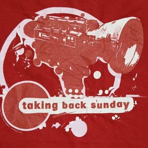 May include: A red t-shirt featuring a white graphic print of a vintage film camera inside a circular frame. Below the camera, a white horizontal banner contains the text "taking back sunday" in red lowercase letters, with additional circular accents.