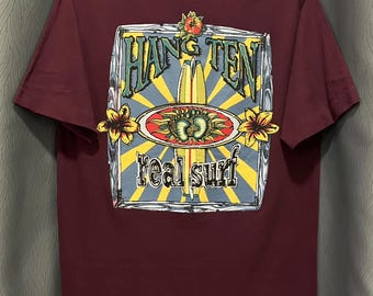 May include: Maroon t-shirt with a graphic on the back featuring the words "Hang Ten" in a turquoise font, two surfboards, and the words "real surf". The design includes yellow, red, and blue accents.
