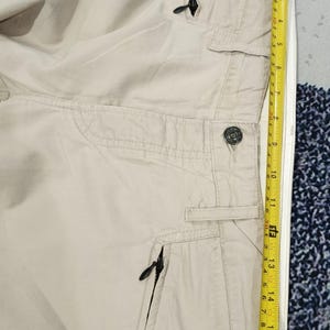 May include: Beige trousers featuring several pockets and black zippers. A yellow tape measure indicates a length of approximately 40 cm.