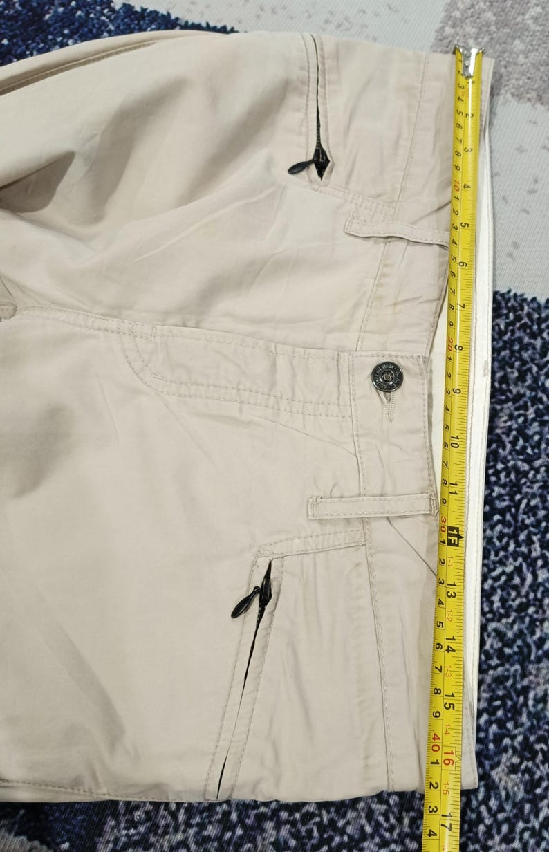 May include: Beige trousers featuring several pockets and black zippers. A yellow tape measure indicates a length of approximately 40 cm.