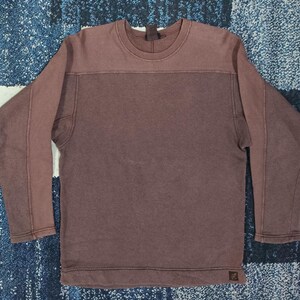 Pode incluir: Uma camisa de manga comprida e gola redonda marrom. A parte superior &eacute; marrom claro, com uma se&ccedil;&atilde;o marrom mais escura abaixo. As mangas t&ecirc;m costuras costuradas. Uma pequena etiqueta &eacute; vis&iacute;vel na parte inferior.
