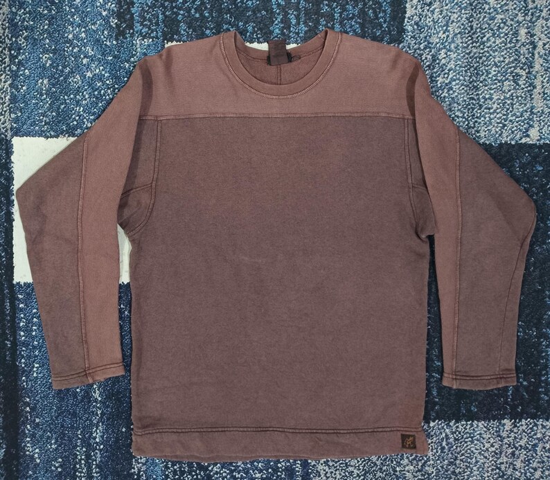 Pode incluir: Uma camisa de manga comprida e gola redonda marrom. A parte superior &eacute; marrom claro, com uma se&ccedil;&atilde;o marrom mais escura abaixo. As mangas t&ecirc;m costuras costuradas. Uma pequena etiqueta &eacute; vis&iacute;vel na parte inferior.