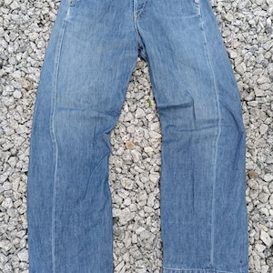 Puede incluir: Vaqueros de mezclilla azul claro con un diseño de pernera ancha. Los vaqueros tienen bragueta con botones, bolsillos delanteros y una etiqueta. El dobladillo de los vaqueros está vuelto. Los vaqueros están sobre una superficie de pequeñas rocas grises.