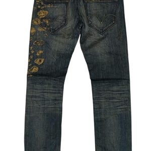 Könnte beinhalten: Dunkelblaue Jeans mit verwaschenem Aussehen. Die Gesäßtaschen zeigen das Levi's-Logo. Das linke Bein hat goldfarbene, abstrakte Designs. Ein weißes Etikett mit Stern und Levi's-Logo befindet sich am Bund.