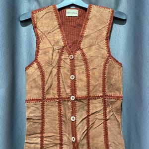 Op de afbeelding: Een vintage bruin suède vest met een gebreide bruine kraag en rug. Het vest heeft een patchwork ontwerp met decoratieve stiksels en witte knopen aan de voorkant. Het label luidt "Christina".