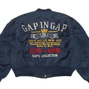 May include: Navy blue bomber jacket with embroidered text and graphics. The back of the jacket features the words "GAP IN GAP" above a crown and banner, along with additional text and circular designs. The jacket has a ribbed collar and cuffs.