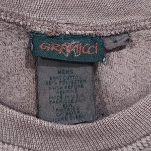 Pode incluir: Close-up de um moletom cinza com textura canelada. A etiqueta da marca Gramicci &eacute; vis&iacute;vel, com o nome da marca em vermelho e verde. A etiqueta tamb&eacute;m inclui a composi&ccedil;&atilde;o do tecido e as instru&ccedil;&otilde;es de cuidado.