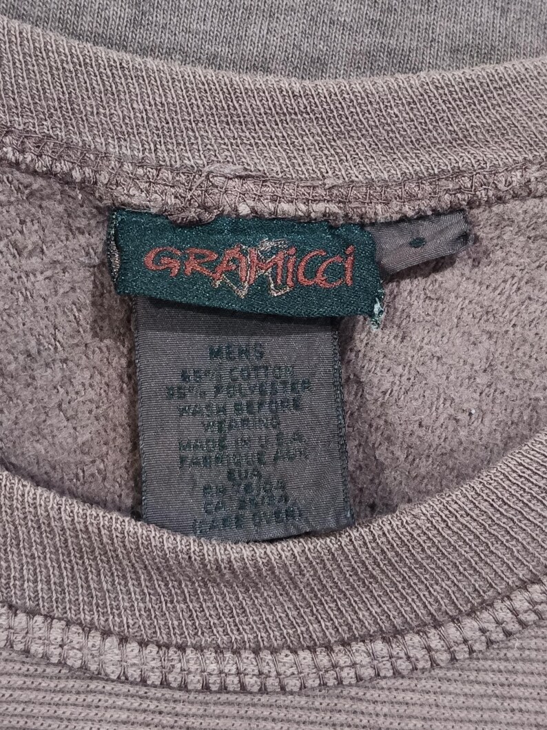 Pode incluir: Close-up de um moletom cinza com textura canelada. A etiqueta da marca Gramicci &eacute; vis&iacute;vel, com o nome da marca em vermelho e verde. A etiqueta tamb&eacute;m inclui a composi&ccedil;&atilde;o do tecido e as instru&ccedil;&otilde;es de cuidado.