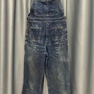 May include: Dark blue denim overalls with a bib front, two chest pockets, and button closures on the sides. The overalls have a flared leg design and adjustable straps. The denim has a worn, faded appearance.