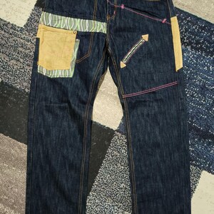 May include: Dark blue denim jeans featuring contrasting tan fabric pockets, green and white wavy patterned panels at the waist and hip, and bright pink stitching accents. The pants include a diagonal zippered pocket and a button fly closure.