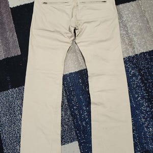 May include: Beige-coloured trousers with a straight leg design, featuring two back pockets with zippers. The trousers are laid flat on a patterned surface, showing the back view of the garment.