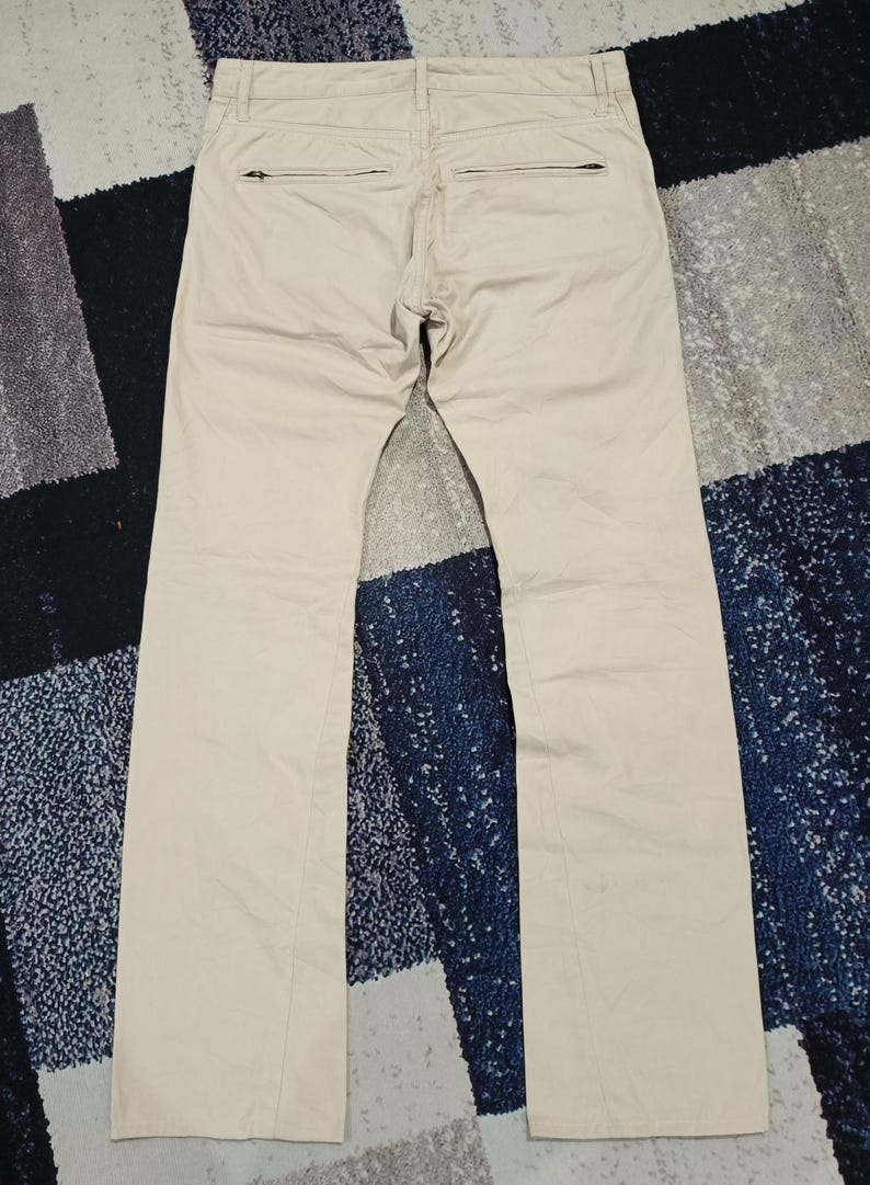 May include: Beige-coloured trousers with a straight leg design, featuring two back pockets with zippers. The trousers are laid flat on a patterned surface, showing the back view of the garment.