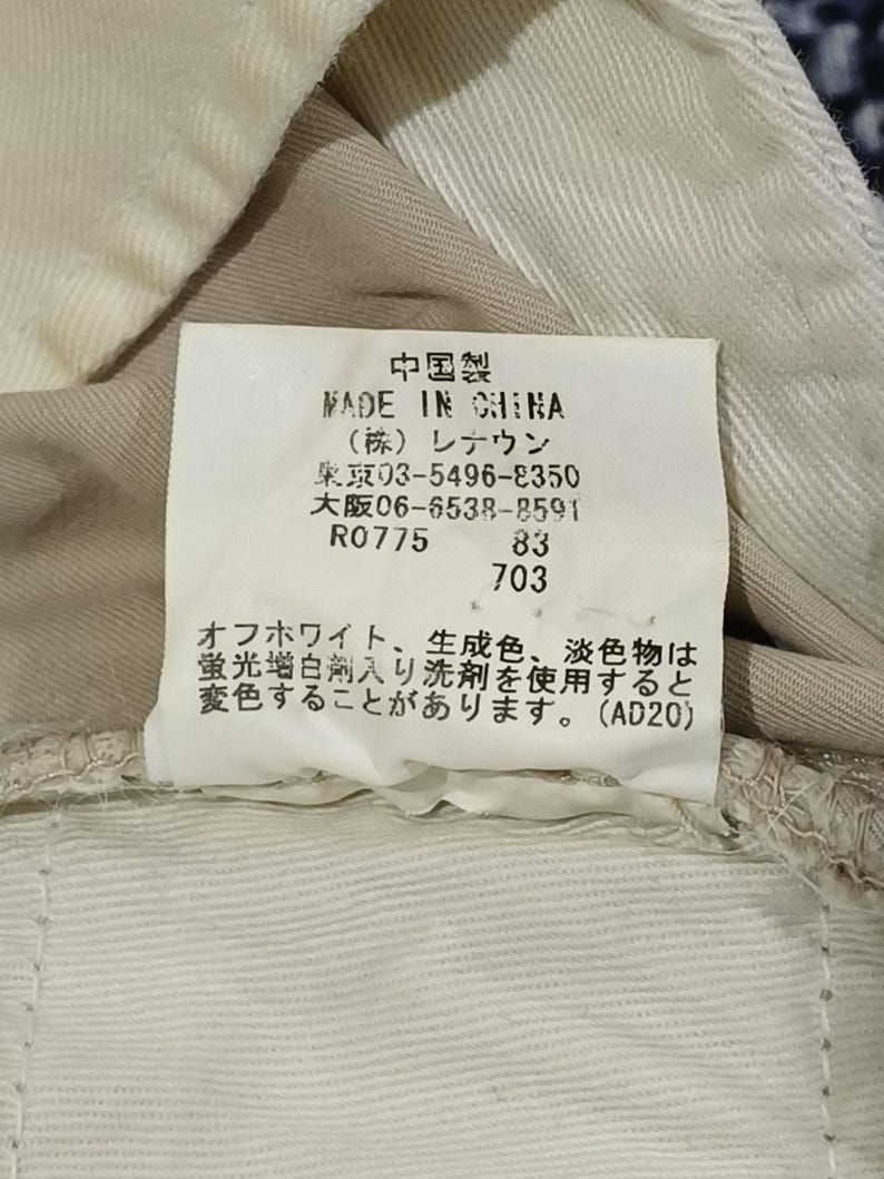May include: A close-up of a tag with Japanese text, including the words "MADE IN CHINA." The tag is attached to beige fabric. The text also includes a warning about using fluorescent whitening agents.