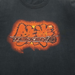 Könnte beinhalten: Ein dunkelgraues Kurzarm-T-Shirt mit einer großen, verwaschenen orange-gelben Grafik auf der Brust. Die Grafik zeigt den Schriftzug TEKKEN 6 in einer stilisierten, fetten Schriftart mit einem feurigen, texturierten Hintergrundeffekt um den Text.