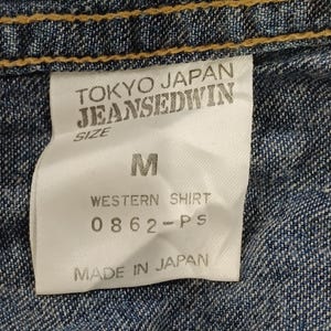 Pode incluir: Close-up de uma etiqueta de camisa de ganga. A etiqueta diz "TOKYO JAPAN JEANSEDWIN" com o tamanho "M". Abaixo, diz "WESTERN SHIRT 0862-PS MADE IN JAPAN". A camisa &eacute; de ganga de lavagem m&eacute;dia com costuras amarelas.