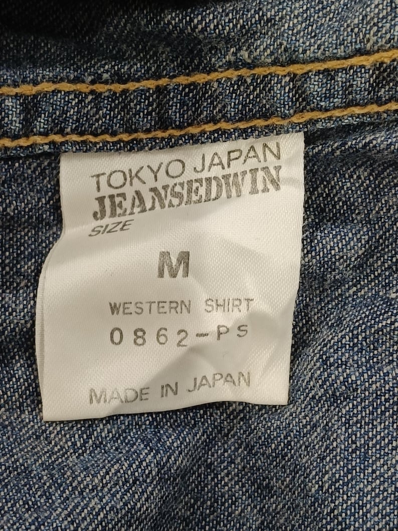 Pode incluir: Close-up de uma etiqueta de camisa de ganga. A etiqueta diz "TOKYO JAPAN JEANSEDWIN" com o tamanho "M". Abaixo, diz "WESTERN SHIRT 0862-PS MADE IN JAPAN". A camisa &eacute; de ganga de lavagem m&eacute;dia com costuras amarelas.