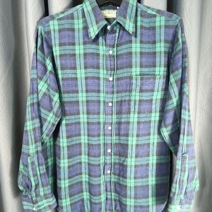 Pode incluir: Uma camisa de flanela de manga comprida com botões em um padrão xadrez de verde-azulado, azul marinho e preto. A camisa tem gola, bolso no peito e punhos abotoados. A etiqueta da marca "LL Bean" é visível.
