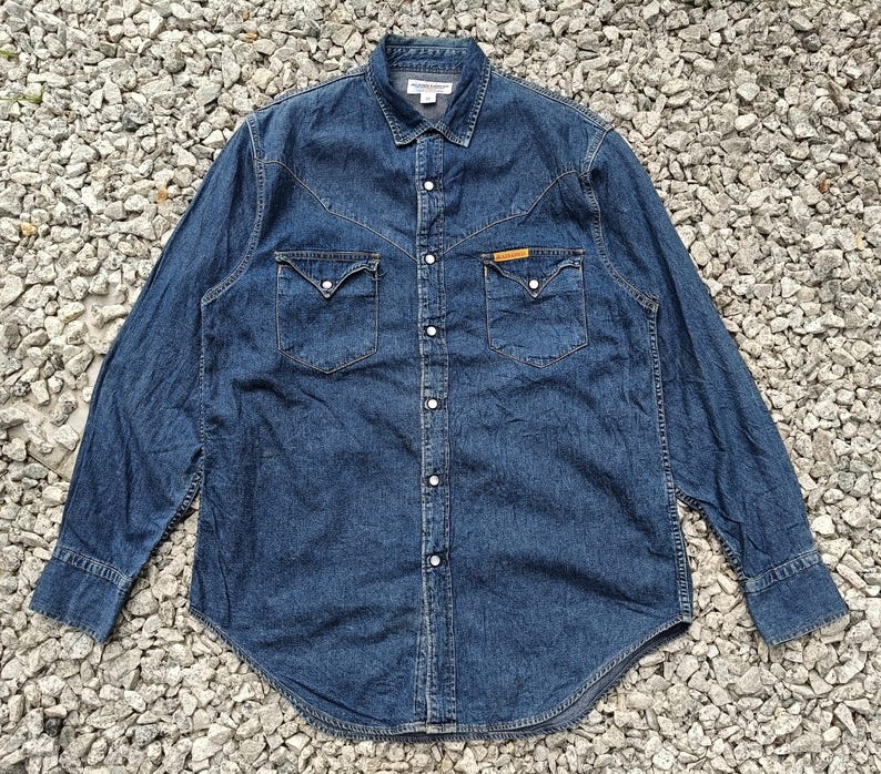 Pode incluir: Camisa de ganga de manga comprida em lavagem azul m&eacute;dia. A camisa tem uma frente abotoada, dois bolsos no peito e uma gola cl&aacute;ssica. A camisa tem um jugo de estilo ocidental e &eacute; feita de tecido de ganga dur&aacute;vel.