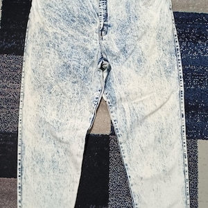 Könnte beinhalten: Hoch taillierte Jeans aus Denim mit einem hellblauen und weißen Acid-Wash-Muster. Die Hose verfügt über einen Knopf- und Reißverschluss, zwei Vordertaschen und einen klassischen geradlinigen Schnitt mit sichtbaren Nähten an den Beinen und am Bund.