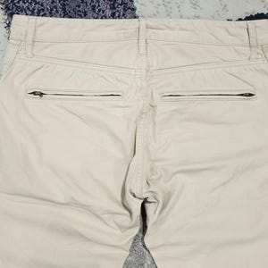 May include: Close-up of the rear of beige trousers. The trousers feature two zipped pockets and a button closure. The fabric looks to be a lightweight material, suitable for warmer weather. The trousers are neatly tailored and appear to be in good condition.