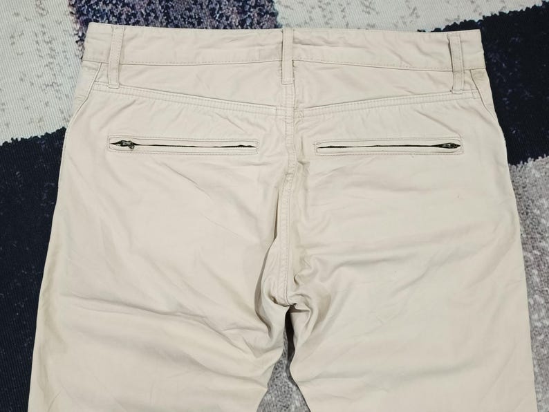 May include: Close-up of the rear of beige trousers. The trousers feature two zipped pockets and a button closure. The fabric looks to be a lightweight material, suitable for warmer weather. The trousers are neatly tailored and appear to be in good condition.