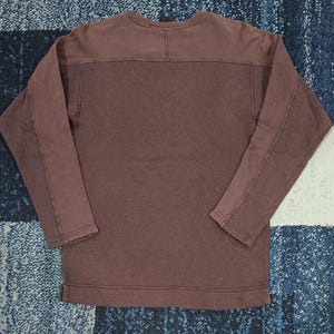 Pode incluir: Uma camisa de manga comprida com gola redonda em um tom marrom suave. A pe&ccedil;a apresenta um design de pain&eacute;is nos ombros e ao longo das mangas. O tecido parece ser um material macio e texturizado.