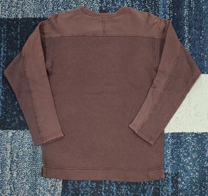 Pode incluir: Uma camisa de manga comprida com gola redonda em um tom marrom suave. A pe&ccedil;a apresenta um design de pain&eacute;is nos ombros e ao longo das mangas. O tecido parece ser um material macio e texturizado.