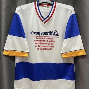 May include: White, blue, and yellow vintage Le Coq Sportif jersey. The shirt has a V-neck with red and blue trim, blue mesh sleeves, and a blue mesh stripe across the midsection. The brand logo and text are printed on the front.