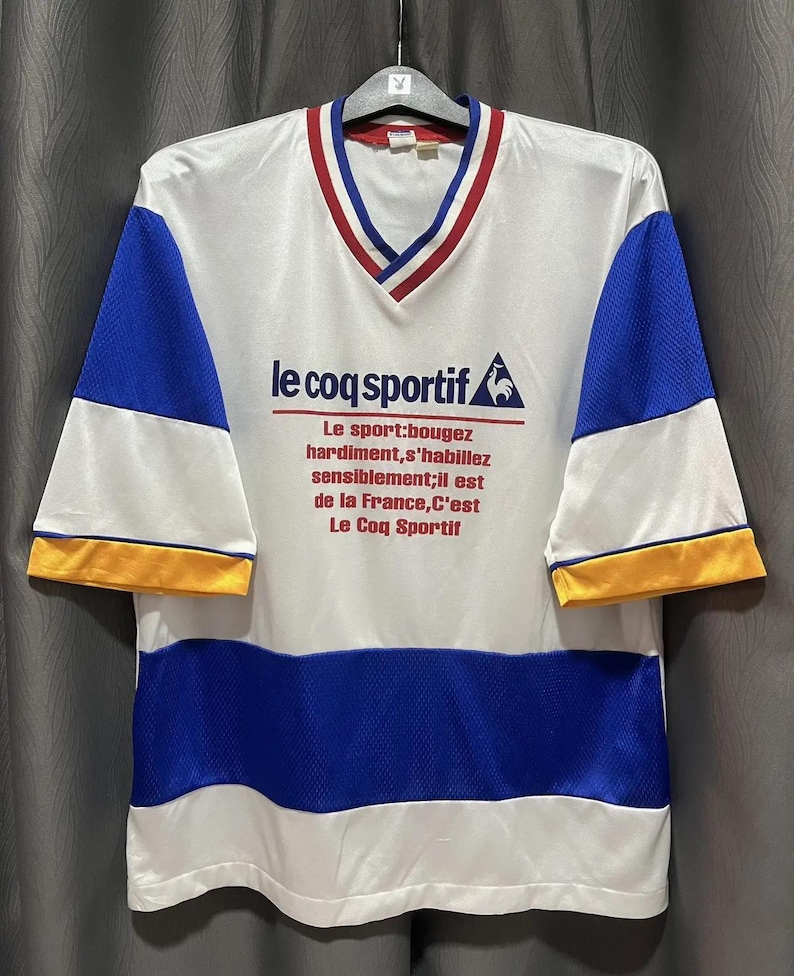May include: White, blue, and yellow vintage Le Coq Sportif jersey. The shirt has a V-neck with red and blue trim, blue mesh sleeves, and a blue mesh stripe across the midsection. The brand logo and text are printed on the front.