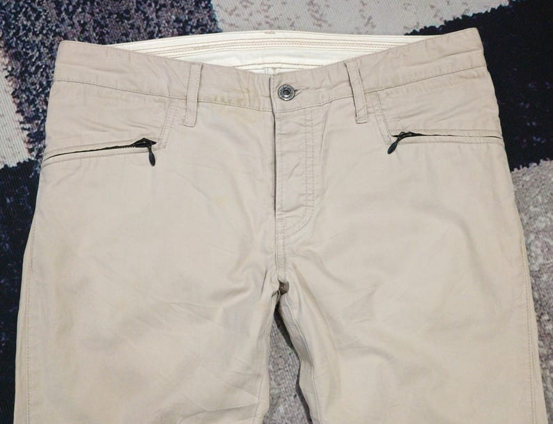 May include: Beige trousers featuring two zipped pockets on the front. The trousers have a button fastening and belt loops. The fabric looks to be a lightweight material, ideal for warmer weather. The trousers are neatly folded, highlighting their design.
