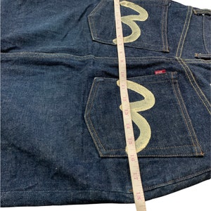 Pu&ograve; includere: Jeans in denim blu scuro con un logo ricamato bianco sulle tasche posteriori. Il logo &egrave; una lettera "M" stilizzata con un nastro di misurazione accanto.