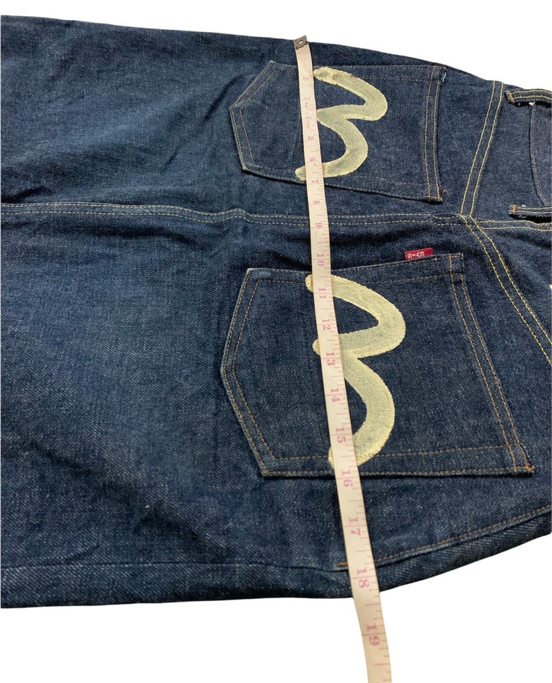 Pu&ograve; includere: Jeans in denim blu scuro con un logo ricamato bianco sulle tasche posteriori. Il logo &egrave; una lettera "M" stilizzata con un nastro di misurazione accanto.