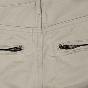 May include: Close-up of the rear of beige trousers, featuring two black zipped pockets. The stitching and fabric texture are visible, highlighting the garment's construction. The zipper pulls are black.