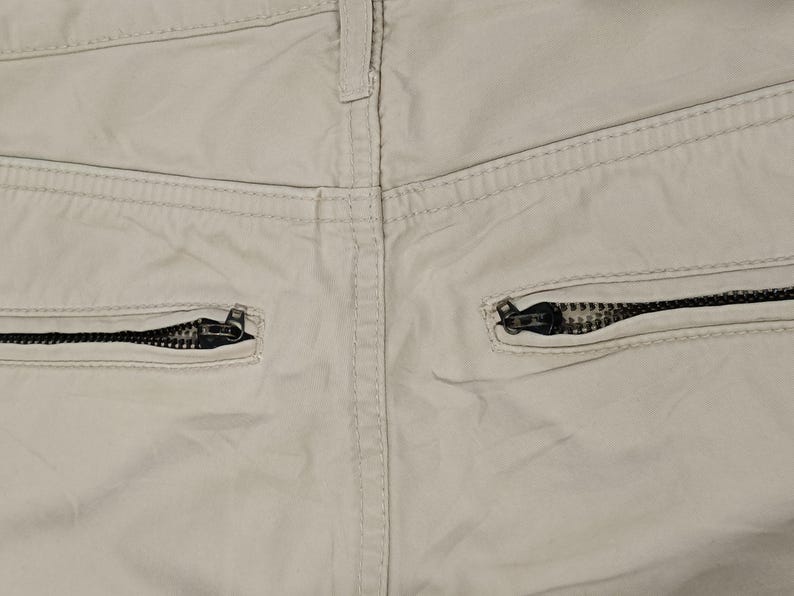 May include: Close-up of the rear of beige trousers, featuring two black zipped pockets. The stitching and fabric texture are visible, highlighting the garment's construction. The zipper pulls are black.