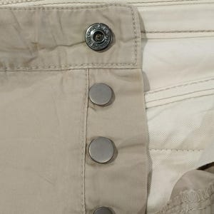 May include: Close-up of beige fabric with a button closure. The closure features a large, decorative button at the top and three smaller, silver-coloured buttons below. The fabric has visible stitching and a layered design.