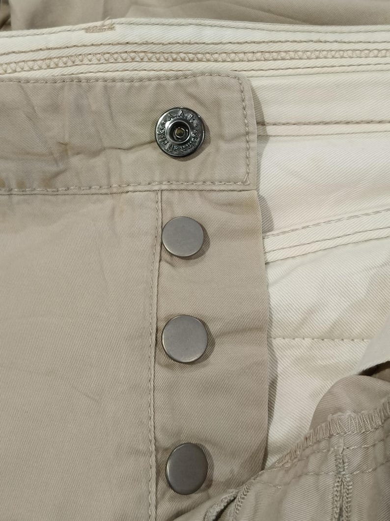 May include: Close-up of beige fabric with a button closure. The closure features a large, decorative button at the top and three smaller, silver-coloured buttons below. The fabric has visible stitching and a layered design.