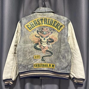Może przedstawiać: Szara i kremowa kurtka uniwersytecka Ghostriders. Z tyłu znajduje się czaszka w kapeluszu kowbojskim, z wężem i płomieniami. Na kurtce widnieje napis "Ghostriders" i "Carlsbad, N.M.".
