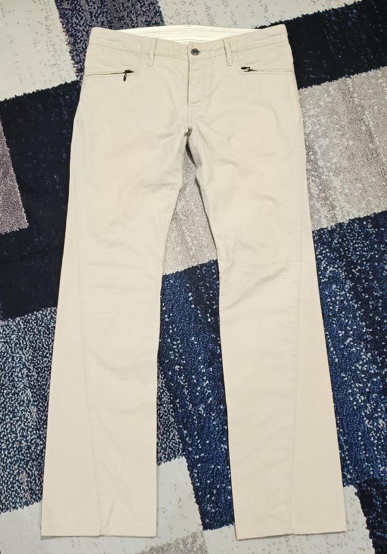 May include: Beige trousers with a classic design. The trousers feature a button and zip closure, along with two zipped front pockets. They are made from a lightweight fabric and have a straight leg cut.