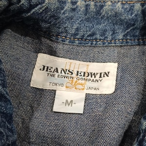 Pode incluir: Close-up de uma camisa jeans azul com uma etiqueta que diz "JEANS EDWIN THE EDWIN COMPANY TOKYO JAPAN -M-". A camisa tem uma textura e costuras cl&aacute;ssicas de ganga.