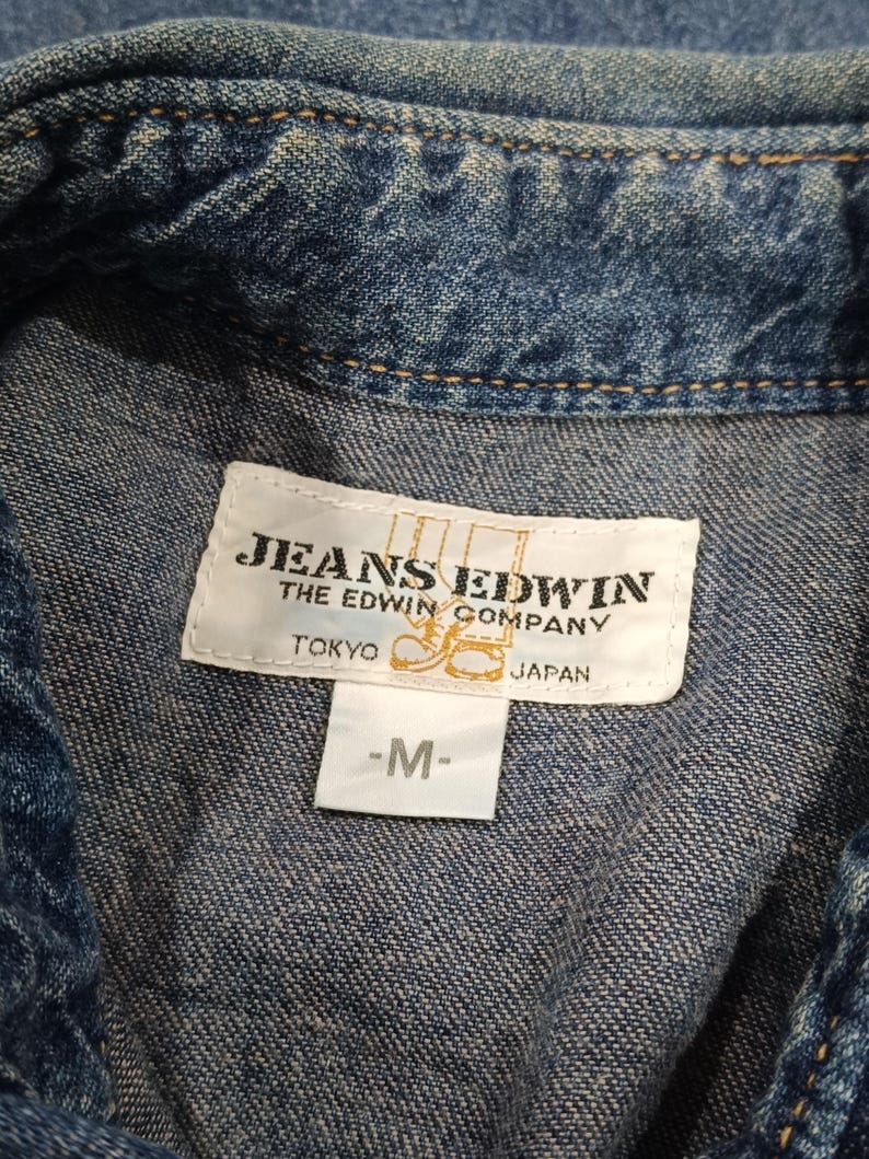 Pode incluir: Close-up de uma camisa jeans azul com uma etiqueta que diz "JEANS EDWIN THE EDWIN COMPANY TOKYO JAPAN -M-". A camisa tem uma textura e costuras cl&aacute;ssicas de ganga.